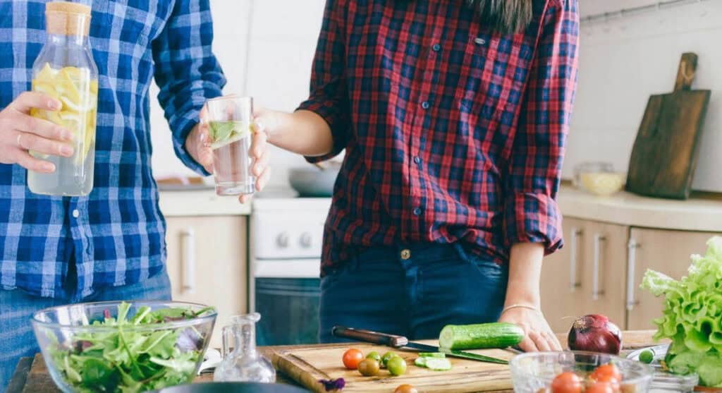 Two people wearing plaid shirts stand in a kitchen, holding glasses with beverages. A countertop in front of them has a cutting board with cucumber slices, a knife, and bowls of salad ingredients.