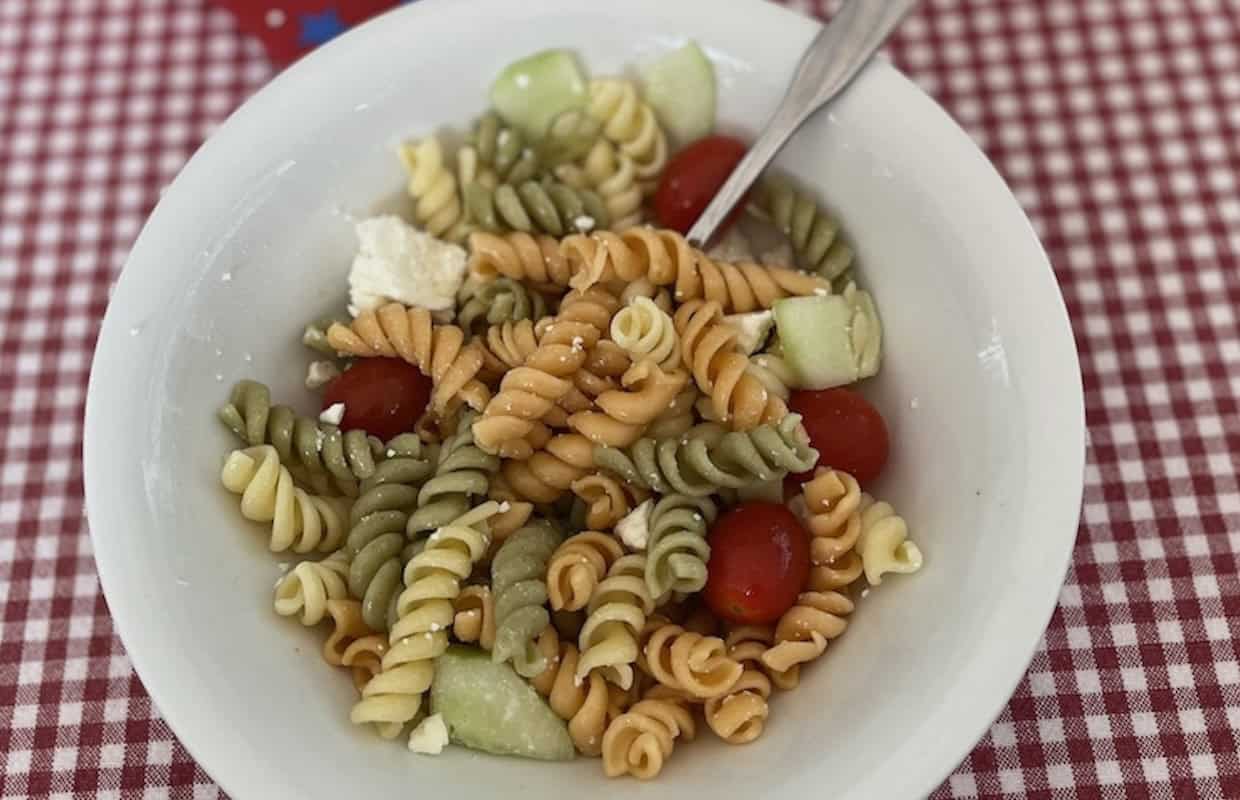 A white bowl filled with tri-color rotini pasta, cherry tomatoes, cucumber chunks, and feta cheese on a red-and-white checkered tablecloth.