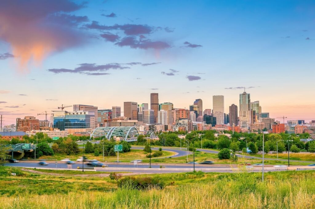 A view of a city skyline with several tall buildings and green spaces in the foreground showcases some highlights when considering things to do in Denver. Roads with moving vehicles are visible, and the sky is partly cloudy during sunset, painting a picturesque urban scene.