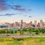 A view of a city skyline with several tall buildings and green spaces in the foreground showcases some highlights when considering things to do in Denver. Roads with moving vehicles are visible, and the sky is partly cloudy during sunset, painting a picturesque urban scene.