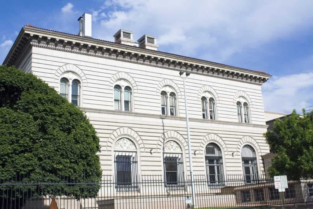 A beige neoclassical building with arched windows, metal bars, and a flat roof stands proudly against a partly cloudy sky. The foreground features a fence and trees, adding to the picturesque scene—one of the charming sights among things to do in Denver.