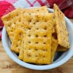 A white bowl filled with seasoned square crackers is placed on a wooden surface, with a red and white checkered cloth in the background.