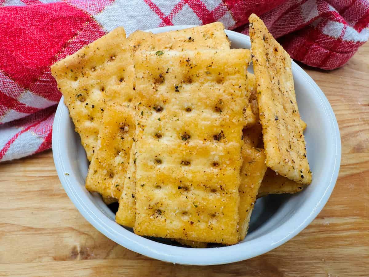 A white bowl filled with seasoned square crackers is placed on a wooden surface, with a red and white checkered cloth in the background.