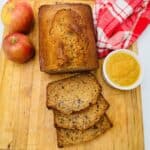 A loaf of applesauce bread with three slices cut, two apples, a bowl of applesauce, and a red and white checkered cloth on a wooden cutting board.