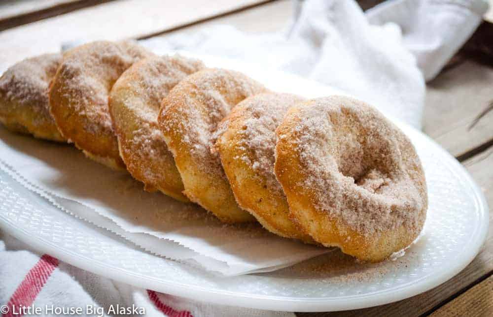 A row of sugar-coated donuts is neatly lined up on a white, oval-shaped plate with a napkin underneath, set on a rustic wooden table.