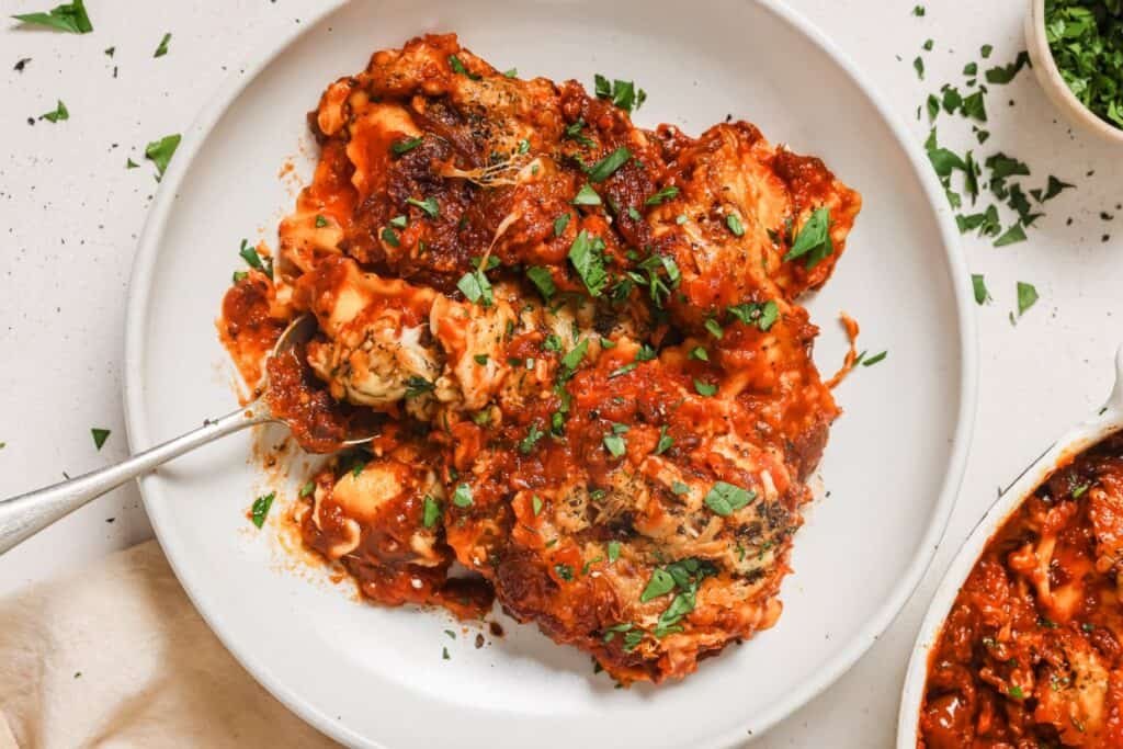 A white plate with a portion of baked ravioli topped with a red tomato sauce and garnished with chopped herbs. A silver fork is positioned on the left side of the plate.