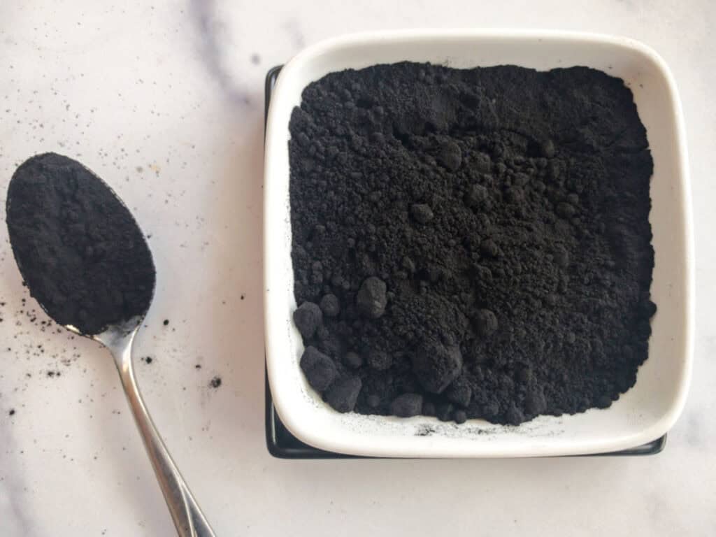 A small white dish filled with powdered activated charcoal sits next to a spoon holding a scoop of the charcoal powder on a marble surface.