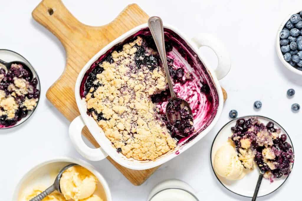 A partially served blueberry crumble in a baking dish with a spoon, surrounded by bowls of the dessert and vanilla ice cream on a wooden board and white countertop.