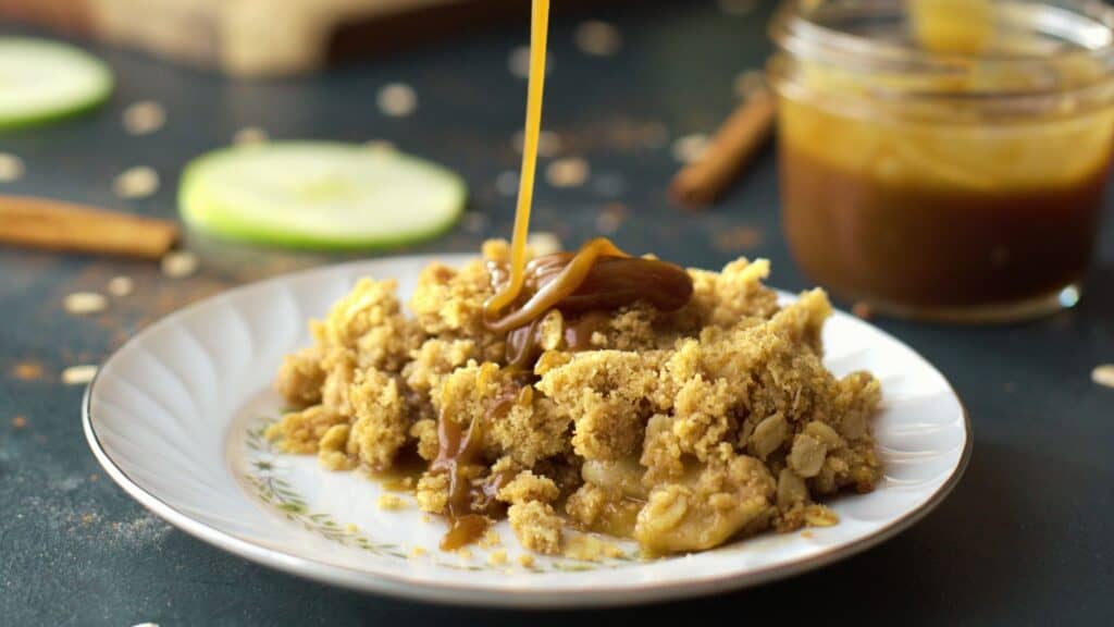 A plate of apple crumble is being drizzled with caramel sauce, with a jar of caramel sauce and a slice of green apple visible in the background.