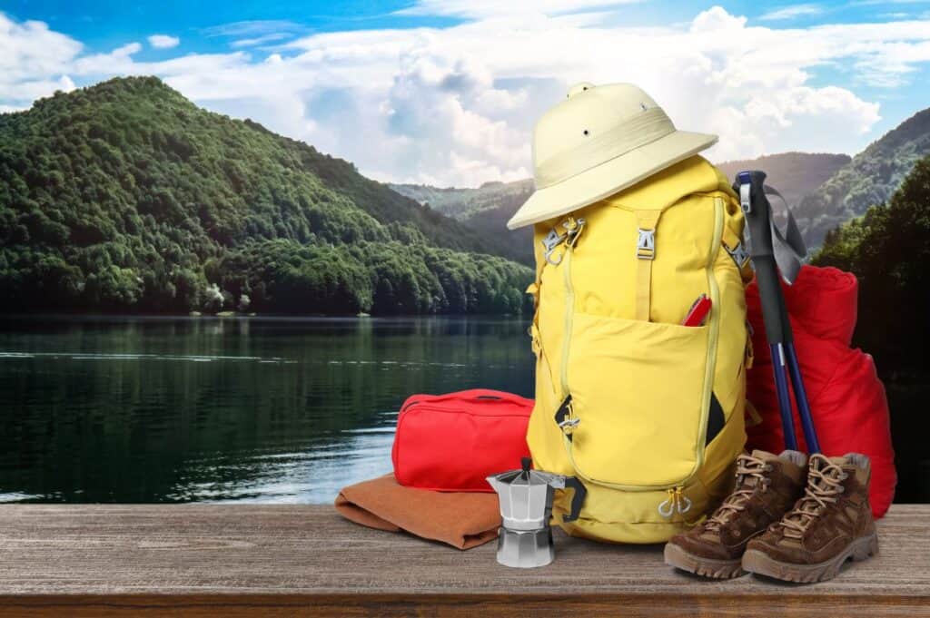 A yellow hiking backpack, boots, hat, walking poles, red bag, and a camp stove on a wooden surface, with a lake and mountainous landscape in the background.