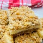 Close-up of crumbly dessert squares with a cream filling, topped with bits of nuts. A red and white checkered cloth is in the background.