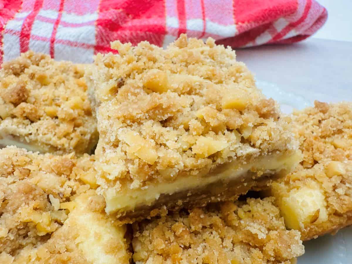 Close-up of crumbly dessert squares with a cream filling, topped with bits of nuts. A red and white checkered cloth is in the background.