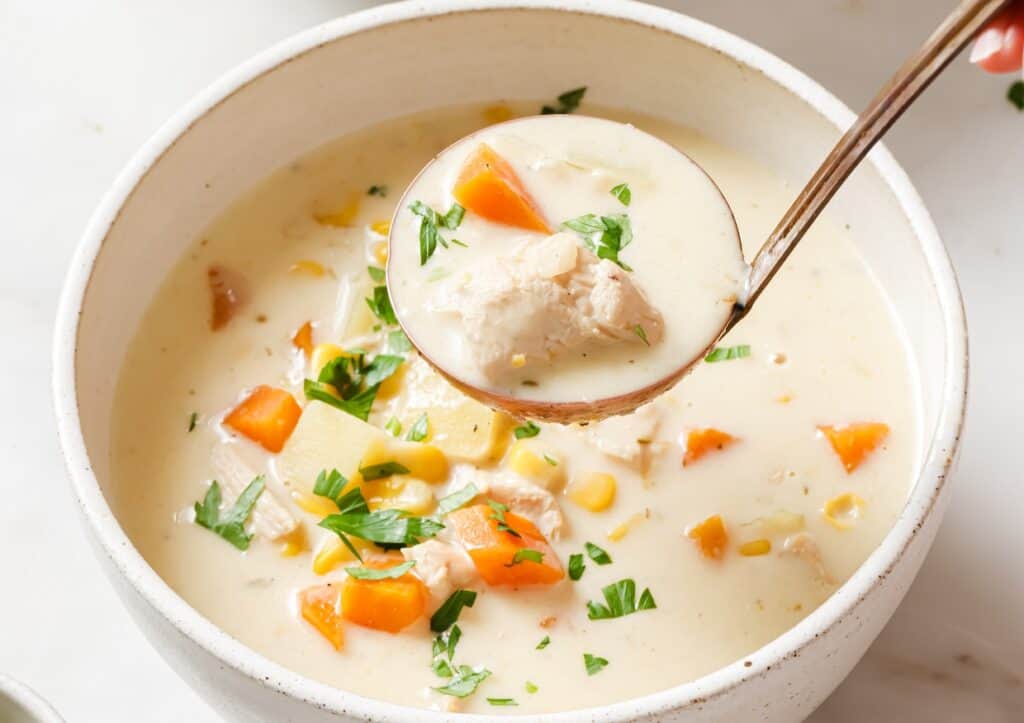 A bowl of creamy soup with vegetables and chunks of chicken, garnished with parsley. A spoon is lifting a portion from the bowl.