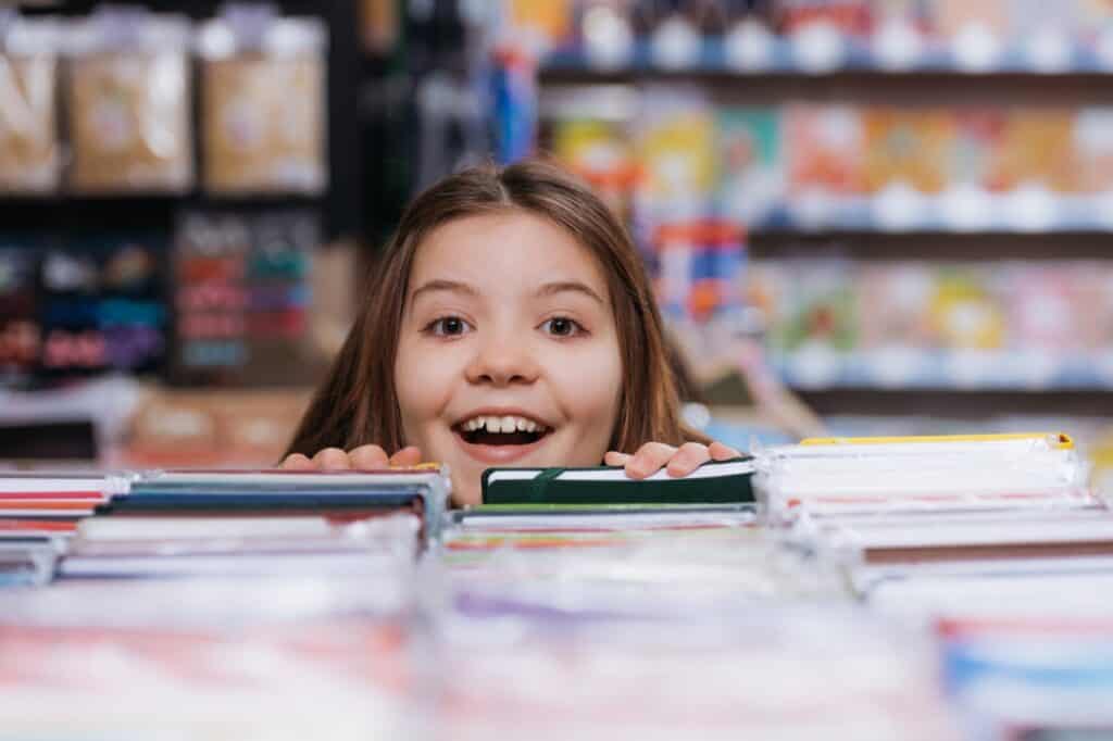 A smiling girl with long hair peeks over a table filled with books in a bookstore. Shelves of more books are visible in the background.