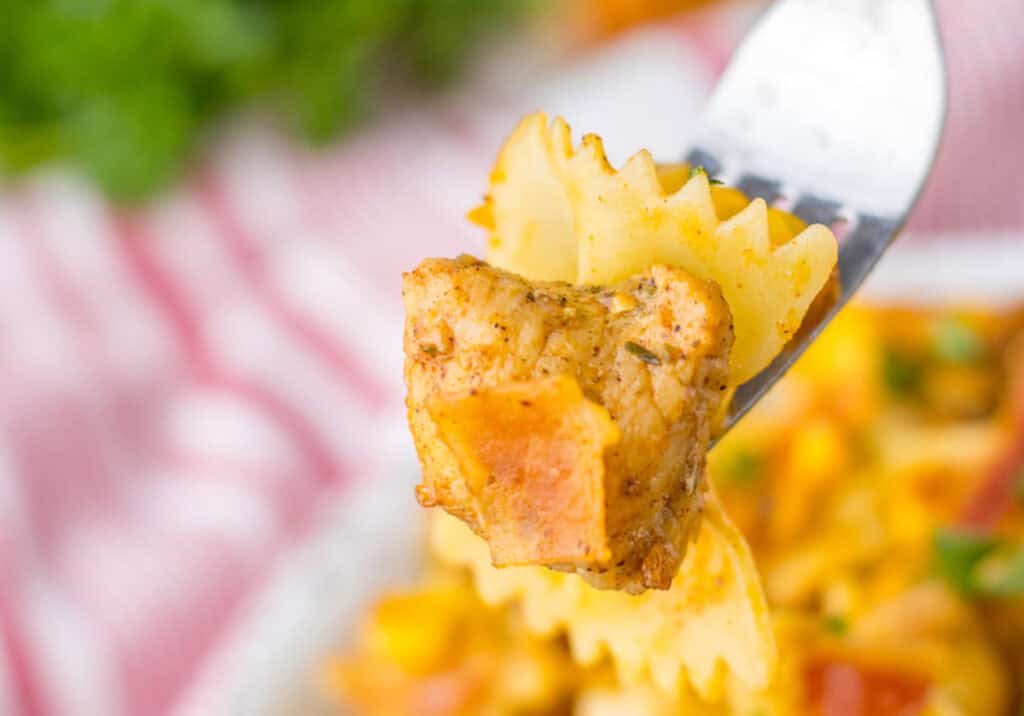 A fork holding a piece of seasoned chicken and farfalle pasta.