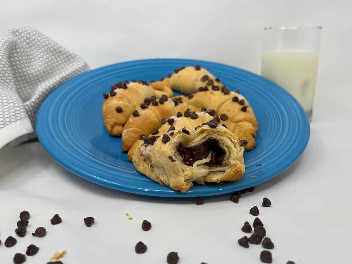 A blue plate with chocolate chip croissants, half-eaten croissant in the front showing chocolate filling, scattered chips on surface, a glass of milk, and a white towel in the background.