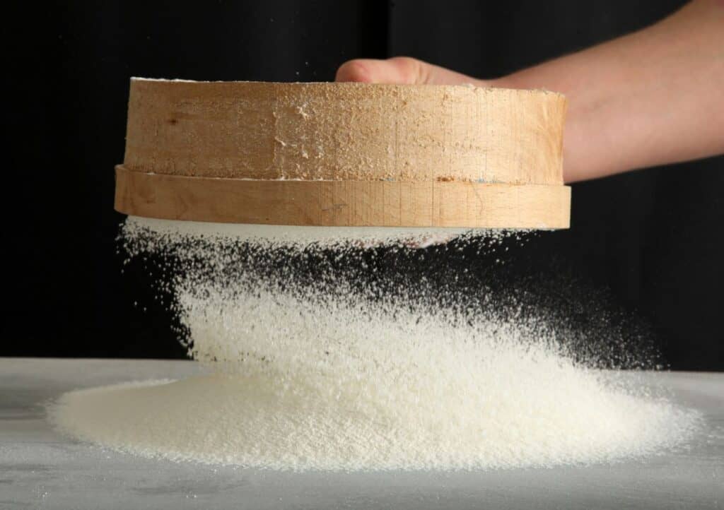 A hand is sifting flour through a round, wooden sieve onto a surface against a dark background.