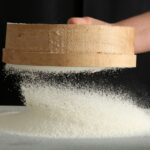 A hand is sifting flour through a round, wooden sieve onto a surface against a dark background.