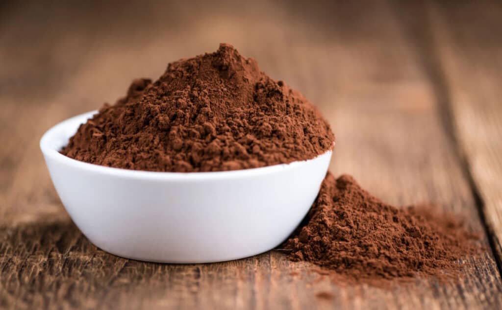 A white bowl filled with cocoa powder, with additional powder spilled on a wooden surface.