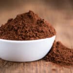 A white bowl filled with cocoa powder, with additional powder spilled on a wooden surface.