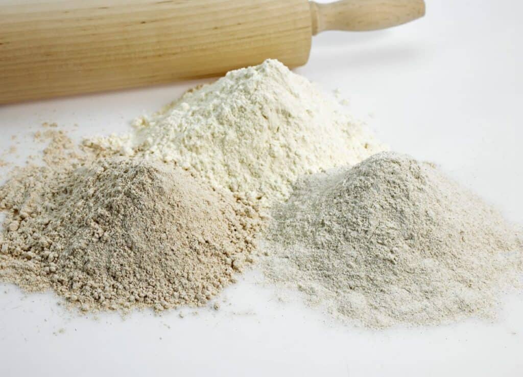 Three piles of flour in different shades of white, brown, and beige are on a white surface with a rolling pin in the background.