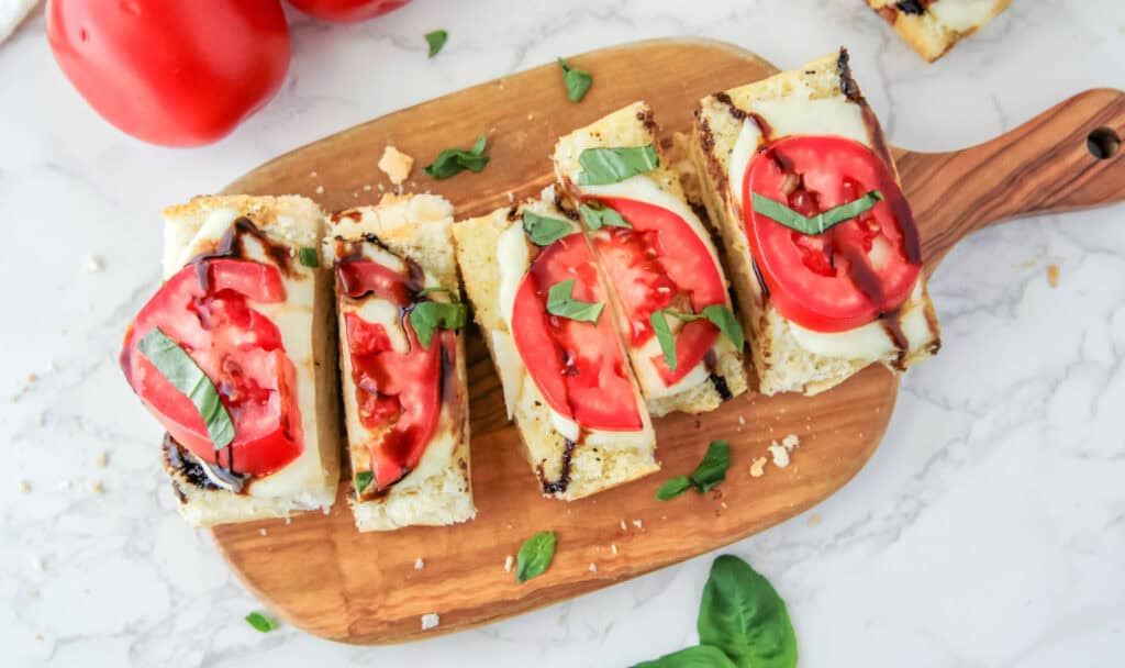 Slices of bread topped with melted cheese, tomato slices, basil leaves, and balsamic glaze, arranged on a wooden board. Fresh tomatoes and basil leaves are nearby.