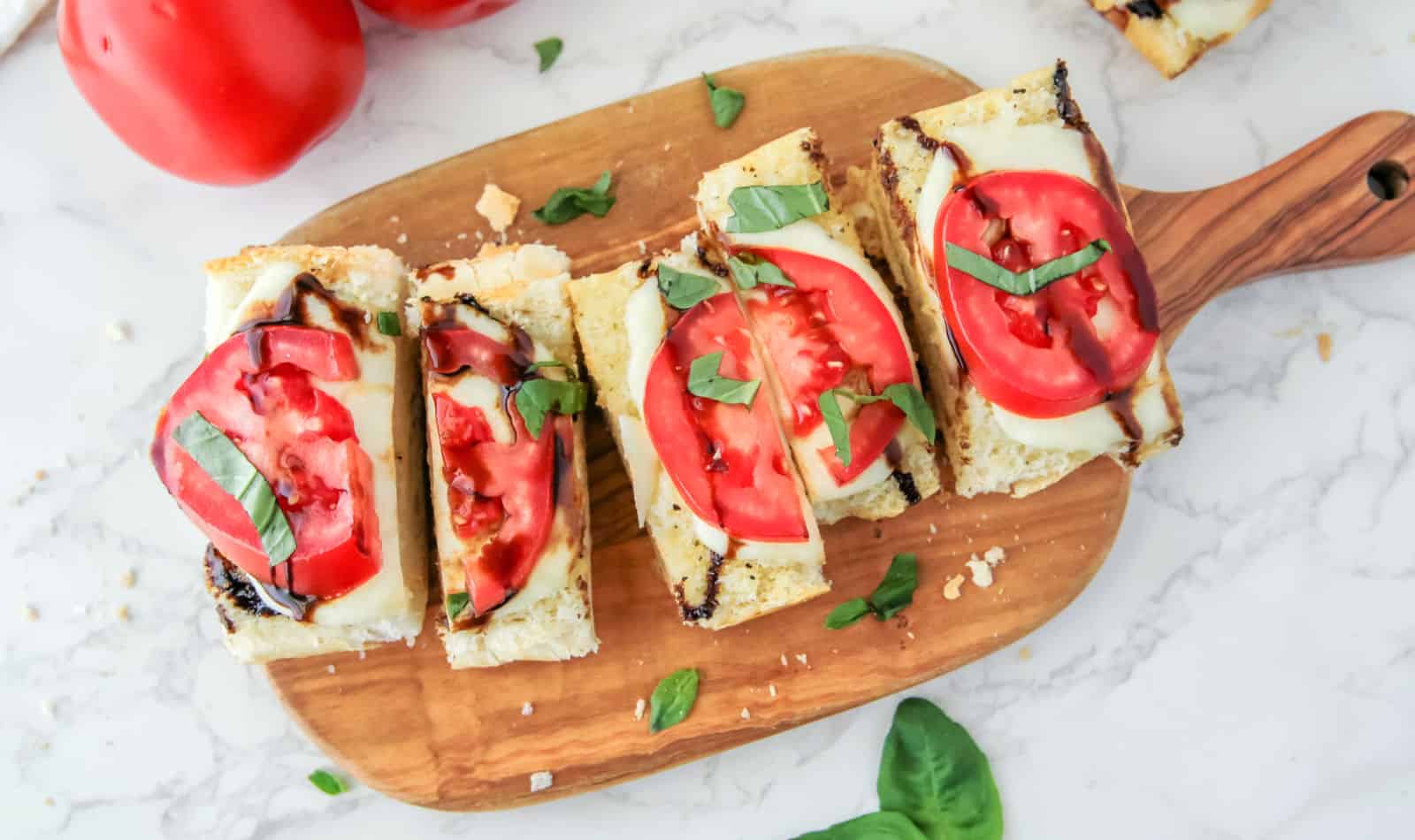 Slices of bread topped with melted cheese, tomato slices, basil leaves, and balsamic glaze, arranged on a wooden board.