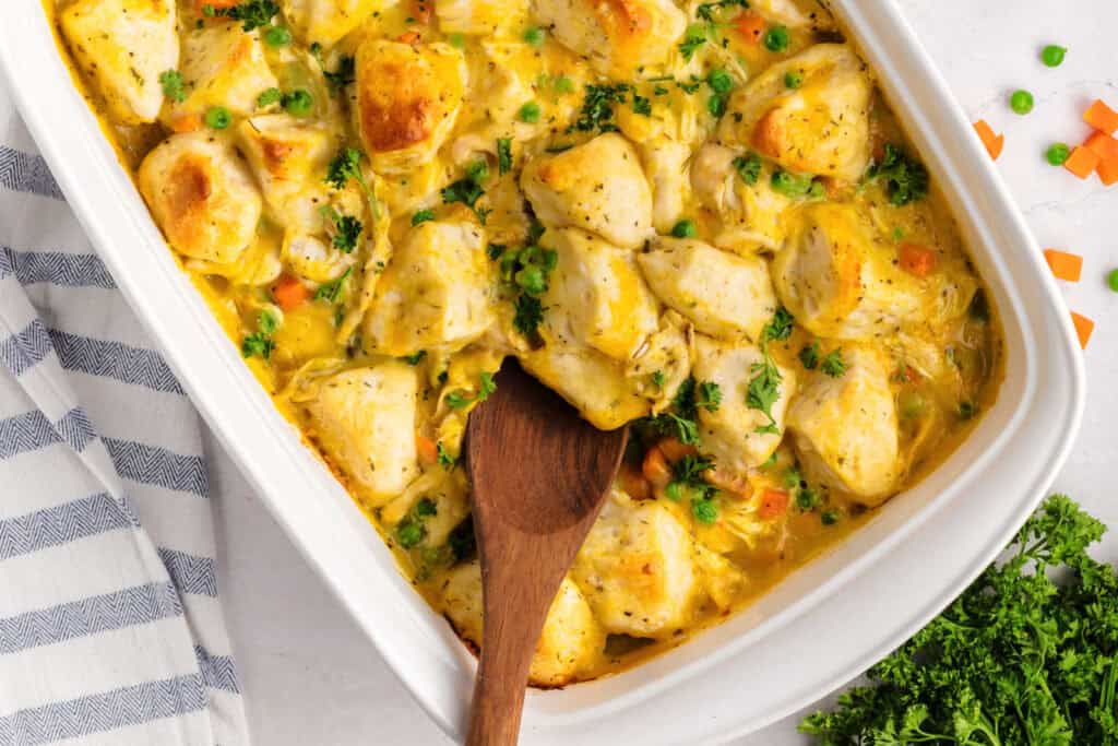 A casserole dish filled with baked chicken, mixed vegetables, and creamy sauce, with a wooden spoon resting inside.