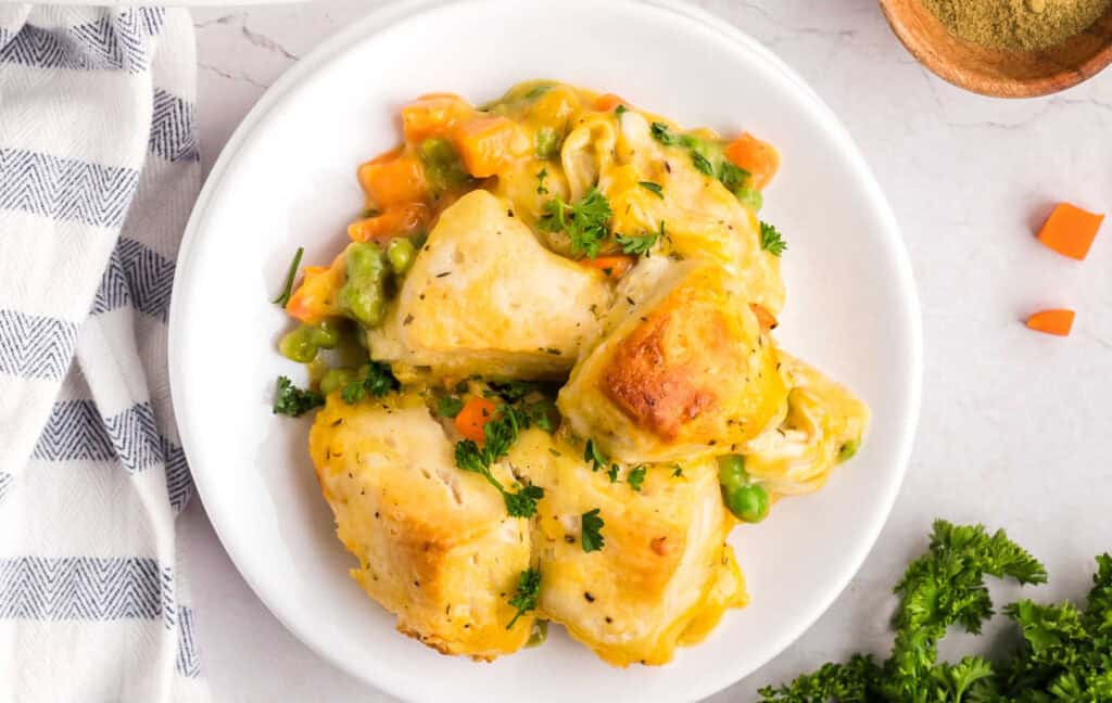 A white plate with a serving of baked chicken pot pie featuring golden-brown biscuits, chicken, peas, carrots, and a creamy sauce. Parsley garnish is visible on top. A napkin and herbs are beside the plate.