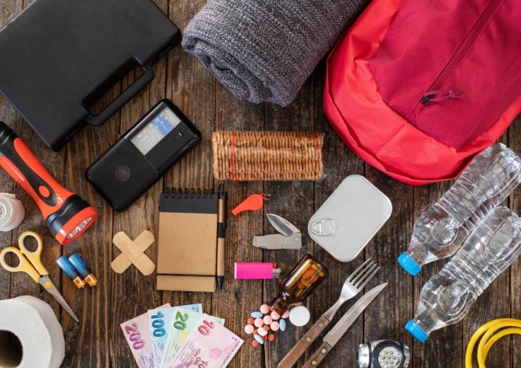 A variety of emergency preparedness items are laid out, including a red backpack, a flashlight, water bottles, first aid supplies, notepad, scissors, and currency notes.