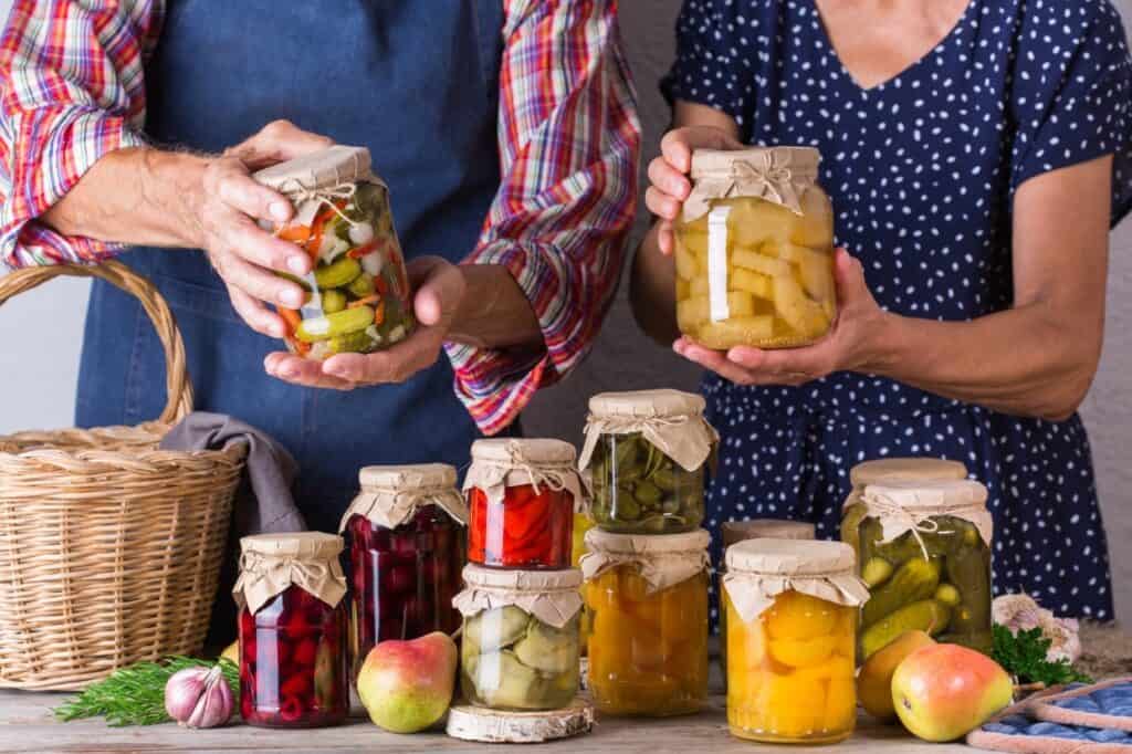 Two people hold jars of preserved food; a variety of other jars are arranged in front of them on a table along with fruits, a wicker basket, and herbs.