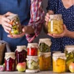 Two people hold jars of preserved food; a variety of other jars are arranged in front of them on a table along with fruits, a wicker basket, and herbs.