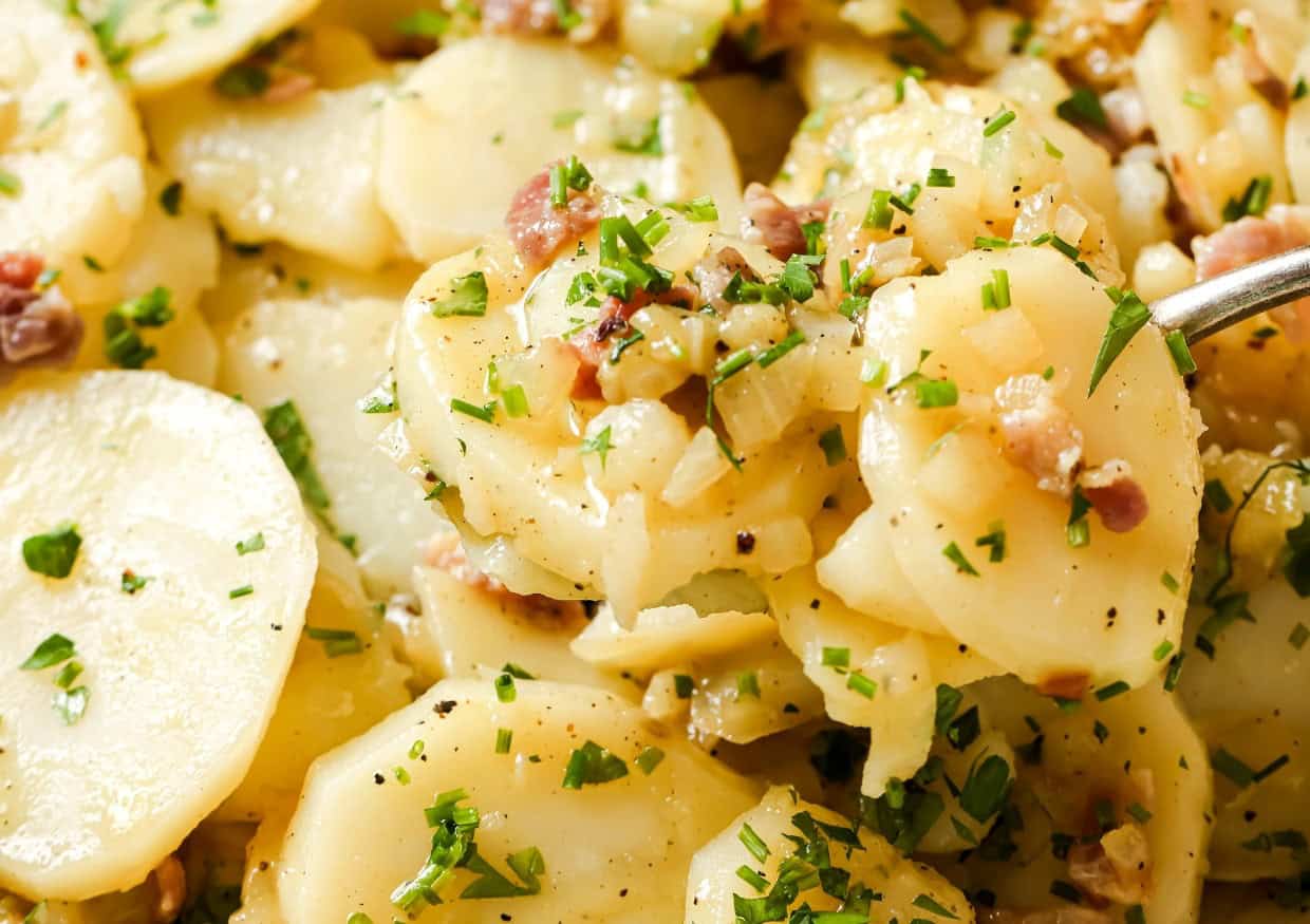 Close-up of a spoon lifting a serving of German potato salad garnished with chopped parsley. The salad contains sliced potatoes, bacon bits, onions, and a light dressing.