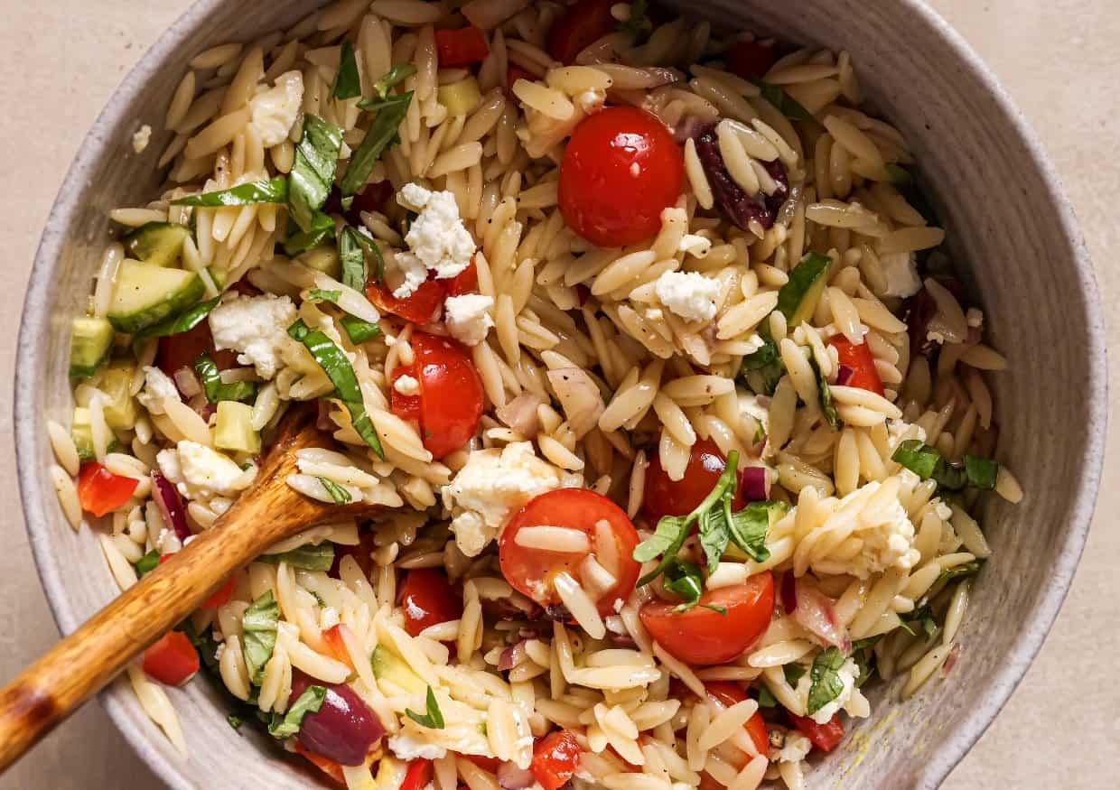 A bowl of orzo pasta salad with chopped tomatoes, olives, cucumbers, red onions, basil, and crumbled feta cheese.