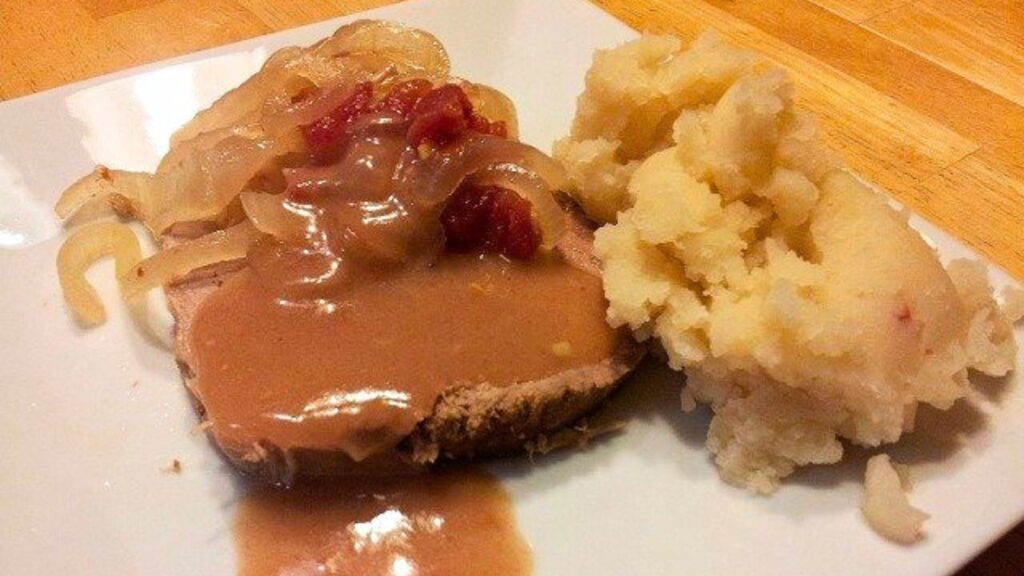 A plate of sliced roast meat topped with gravy, sautéed onions, and a few diced tomatoes, accompanied by a serving of mashed potatoes.