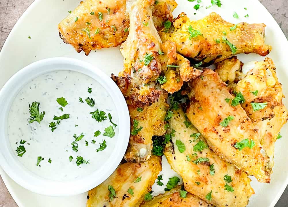 A plate of seasoned chicken wings garnished with chopped parsley, served with a side of ranch dipping sauce in a small white bowl.