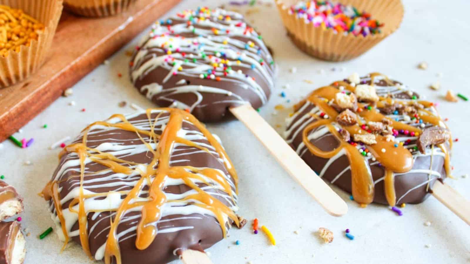 Chocolate-covered apple slices on sticks are decorated with various toppings, including caramel drizzle, sprinkles, and crushed cookies, with more sprinkles in the background.