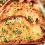 Slices of garlic bread garnished with chopped parsley and sea salt are arranged on a wooden board.