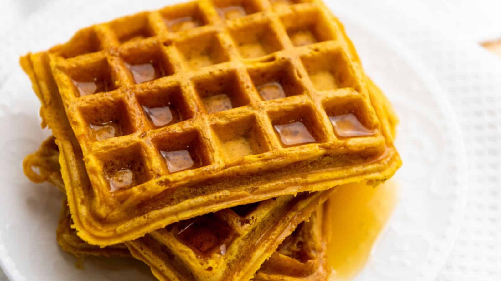 Three golden-brown vegan pumpkin waffles stacked on a white plate, with syrup pooling in their square pockets.