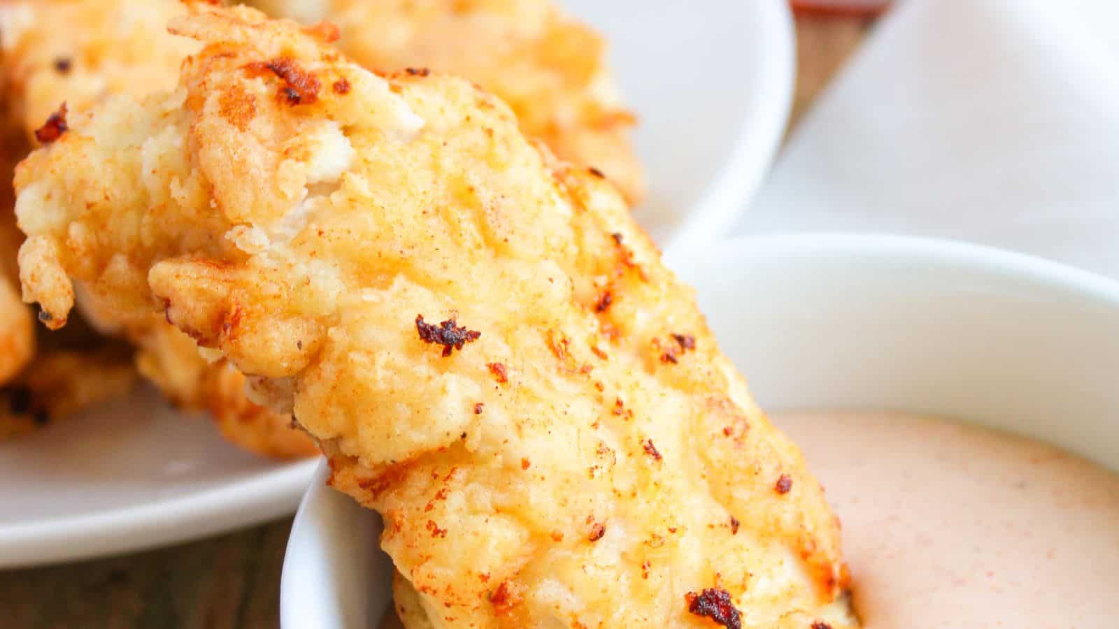 A breaded and golden-brown fried chicken tender being dipped into a white sauce in a bowl, with more chicken tenders in the background.