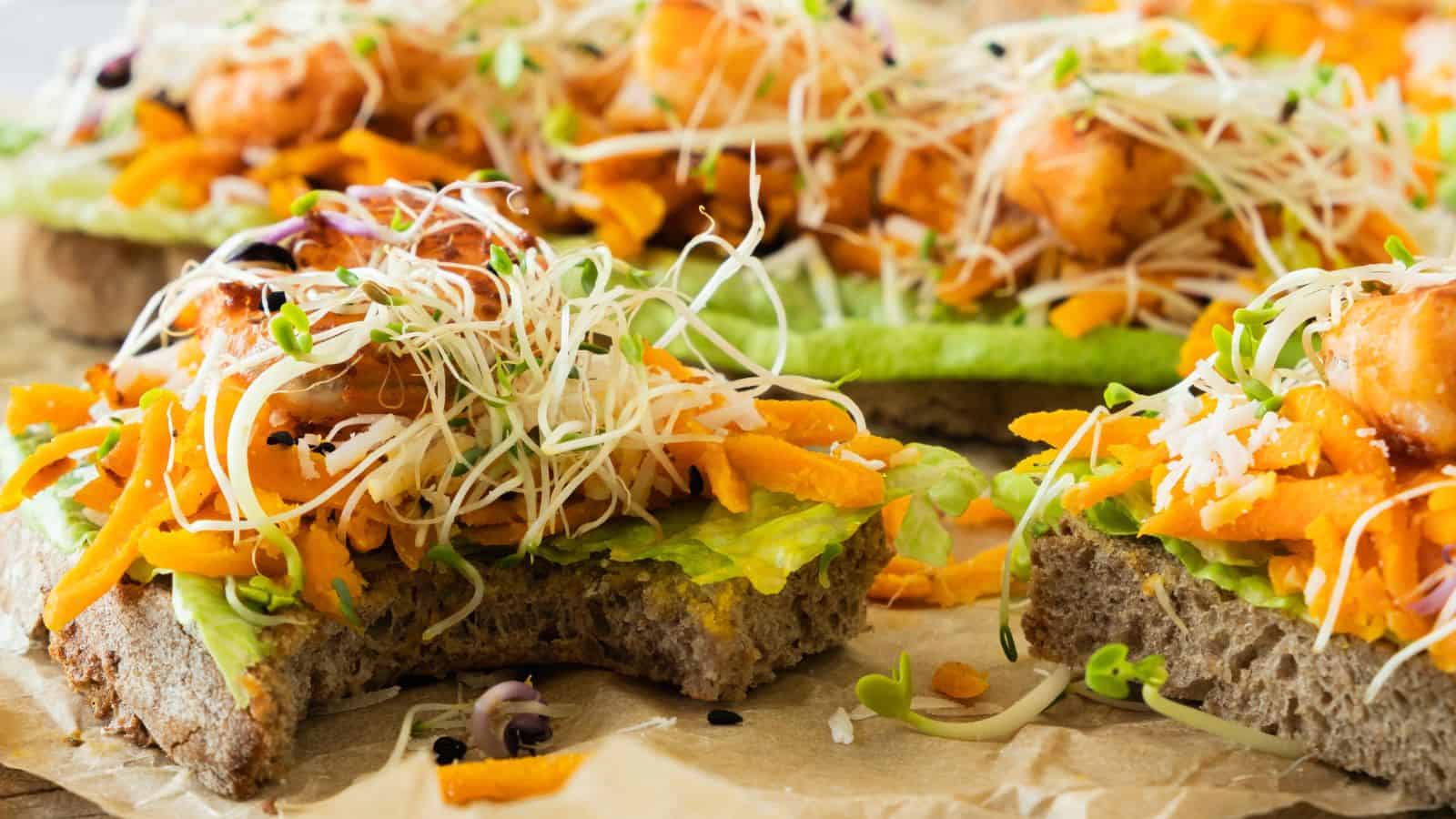 Close-up of open-faced sandwiches topped with avocado, shredded carrot, microgreens, and pieces of cooked salmon on whole-grain bread.