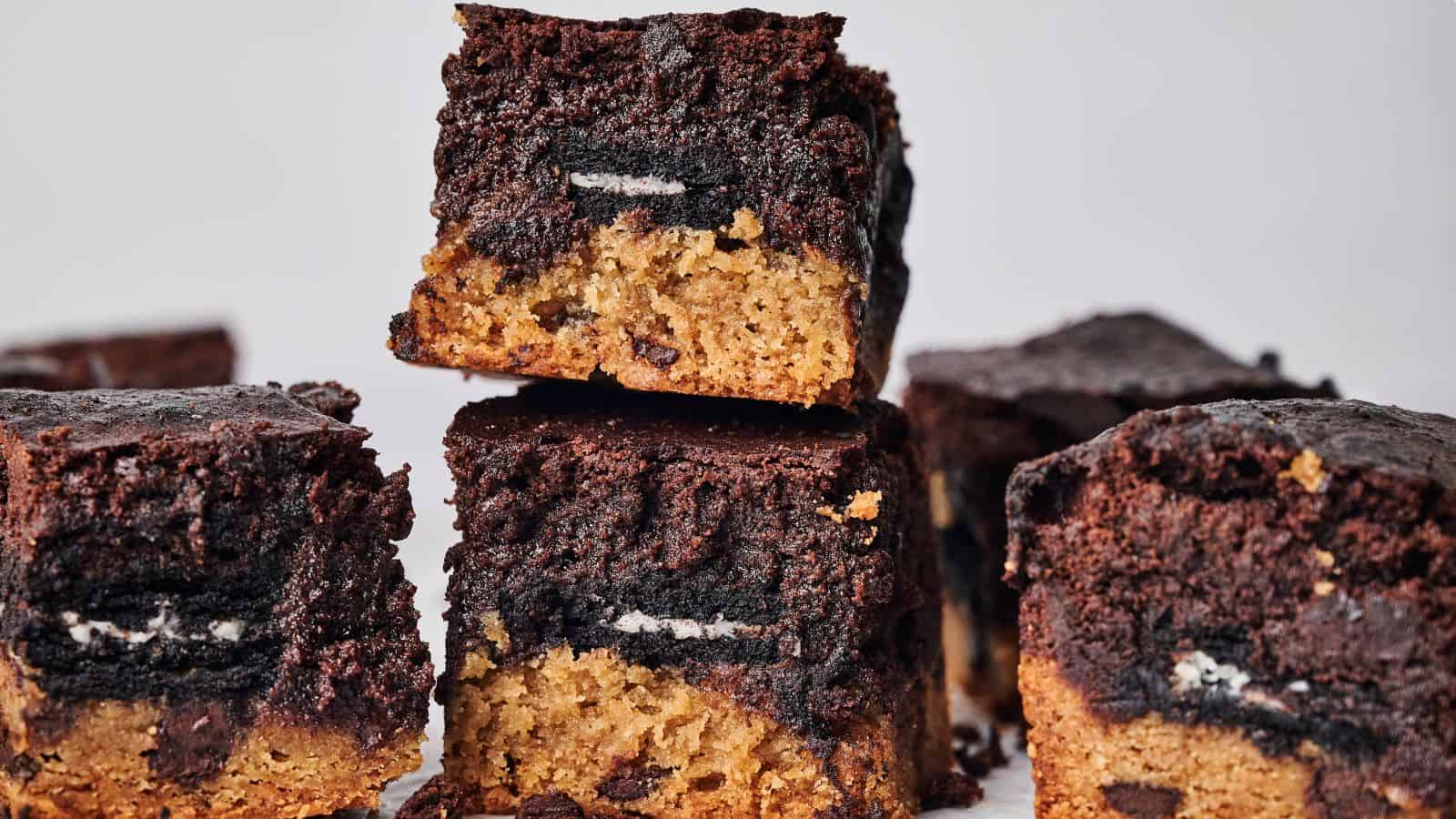 Close-up of multiple chocolate and cookie layered brownies stacked and scattered on a white surface. The brownies feature a brown base with a dark chocolate top layer and a visible Oreo in the middle.