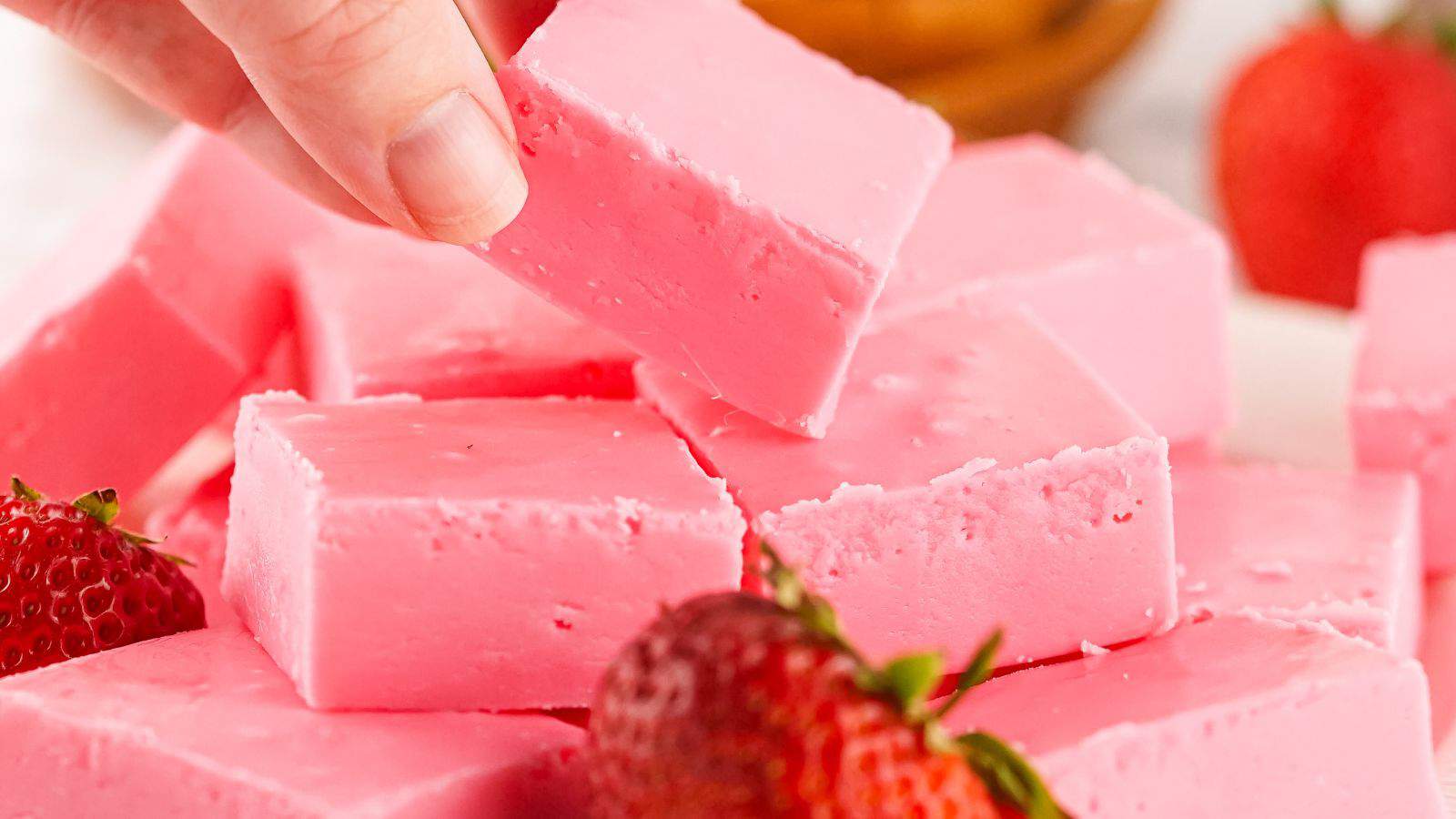 A hand picking up a square piece of pink fudge from a stack, with strawberries in the background.