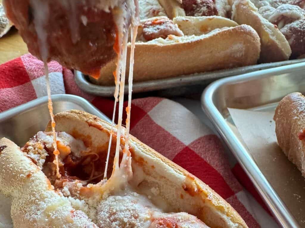 A close-up of a meatball sub with melted cheese being pulled apart, showing strings of cheese between meatballs, on a background of red and white checkered cloth.