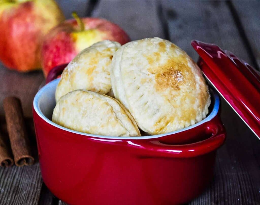 A red dish holds three baked pastry puffs, with a couple of apples and cinnamon sticks in the background on a wooden surface.