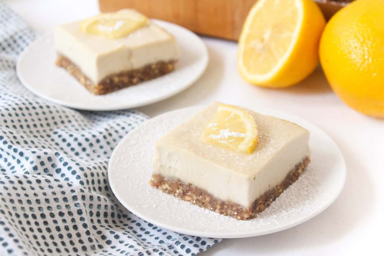 Two plates of lemon bars with a crust and creamy filling, each topped with a lemon slice. Whole lemons and a blue dotted cloth are in the background.