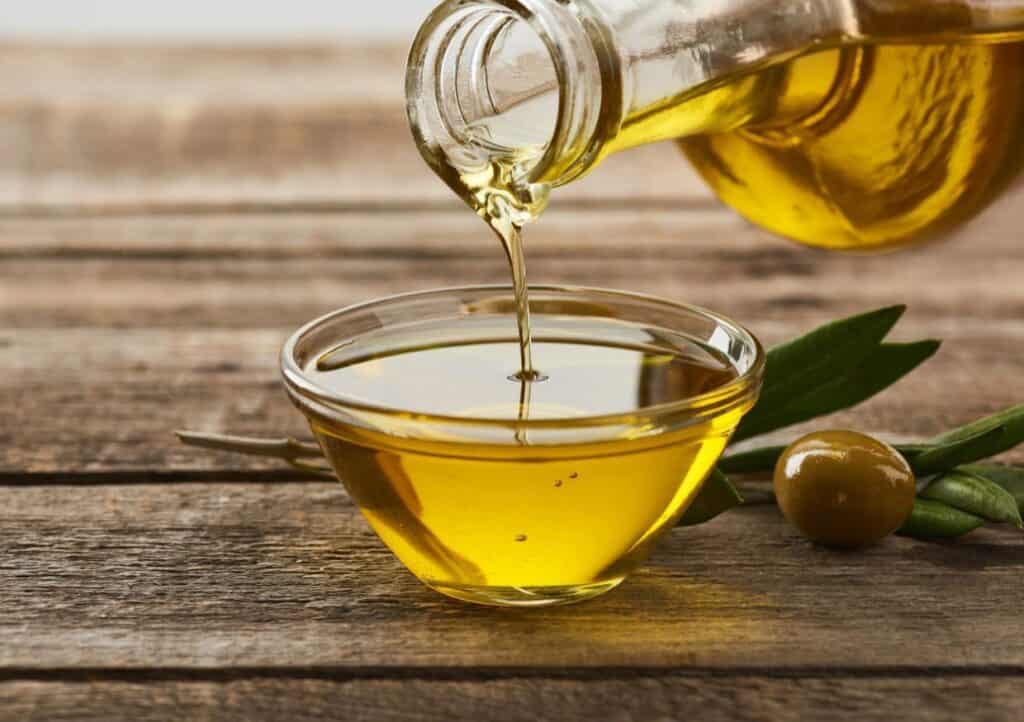 A bottle of olive oil is being poured into a small glass bowl on a wooden surface next to an olive and a twig of olive leaves.