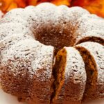 A close-up of a powdered sugar-dusted bundt cake with several slices cut and placed on a white plate. Orange foliage is visible in the background.