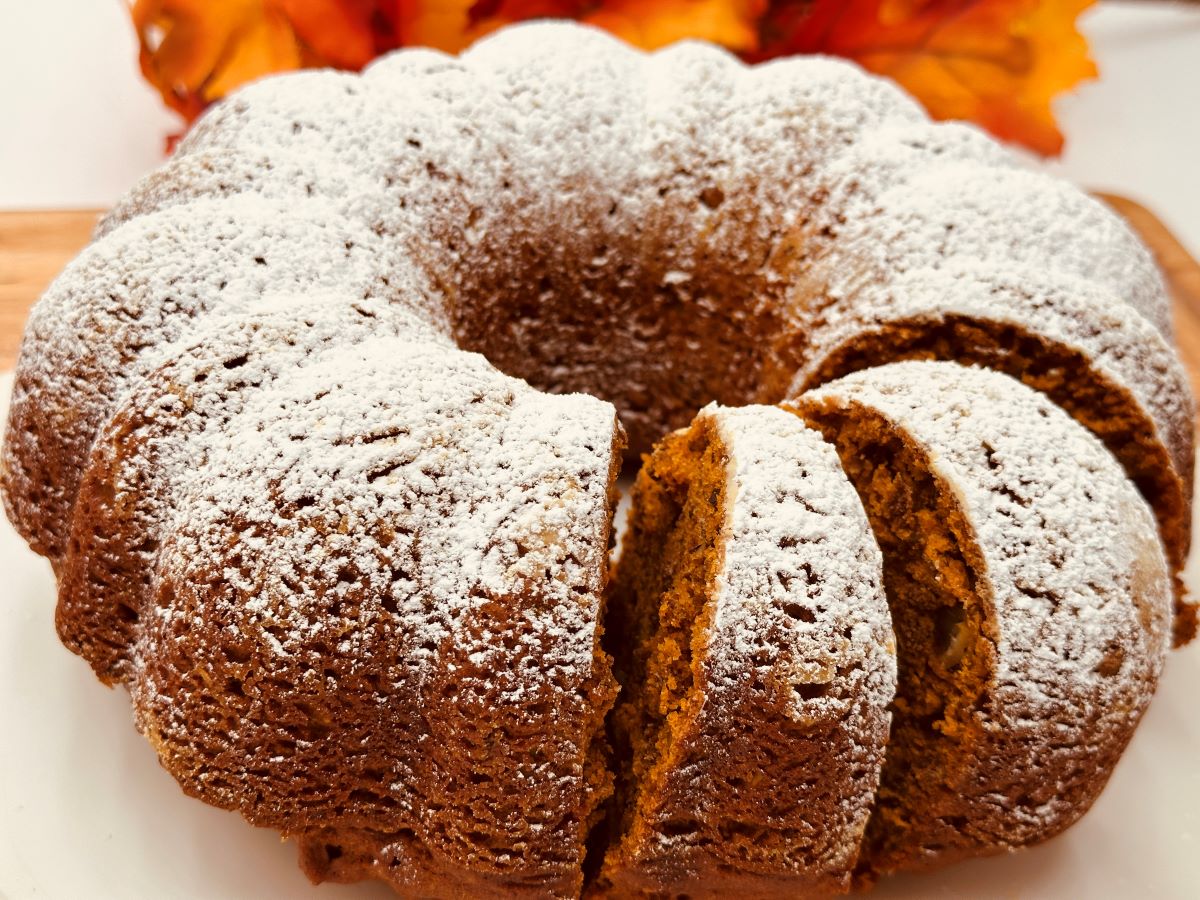 A close-up of a powdered sugar-dusted bundt cake with several slices cut and placed on a white plate. Orange foliage is visible in the background.