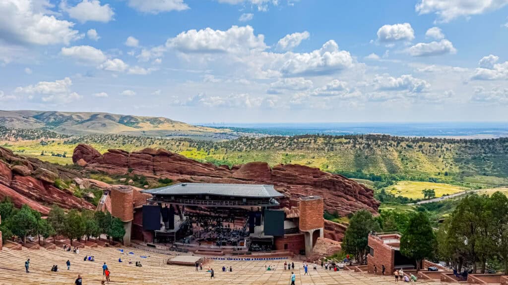 Panoramic view of Red Rocks Amphitheatre with scattered visitors, surrounded by red rock formations and greenery, under a partly cloudy sky. Distant city skyline visible on the horizon.